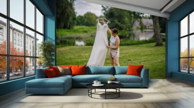 Valmiera, Latvia - August 19, 2023 - A woman in casual attire examines a wedding dress she is holding up in a lush green outdoor setting near a lake. Wall mural