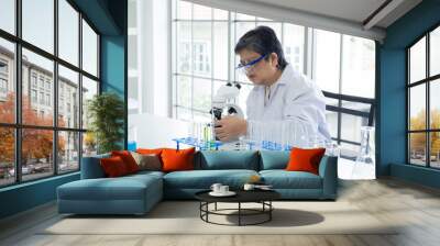 Asian senior female scientist wearing safety goggles and a white lab coat examining chemical samples using a microscope in modern laboratory, various lab equipment and test tubes are arranged on table Wall mural