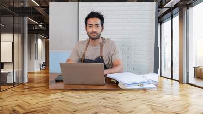 Handsome Indian male restaurant owner sit and looking at camera with confidence. Man employee wearing apron calculate account income and expenses of shop, personal business concept. Wall mural