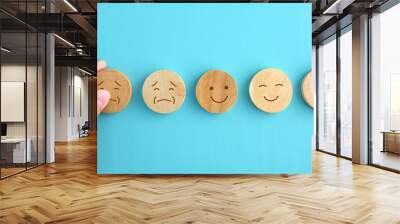 five wooden discs on a blue background, each with a different facial expression. Three discs show negative or neutral emotions, while two feature smiles. A hand is seen picking up one of the smiling Wall mural