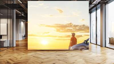 A young man in a shirt enjoys the sunset. A man and his car in a field against the backdrop of a brightly lit sunset sky. Local travel concept. Vertical orientation, space for text. Wall mural