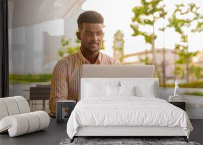 Handsome young African businessman in city using laptop computer at coffee shop Wall mural