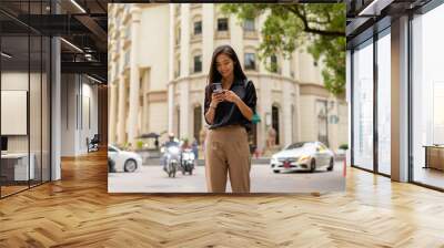 Happy Asian businesswoman outdoors in city street using mobile phone while texting Wall mural