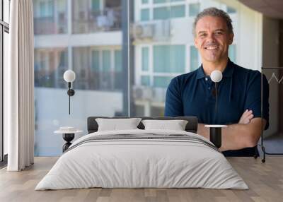 Happy mature handsome Hispanic man smiling with arms crossed by the glass window at home Wall mural