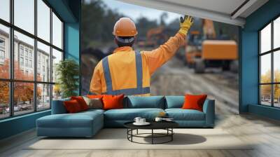 Construction worker in reflective gear and helmet signalling to heavy machinery on a dirt road at a worksite. Wall mural