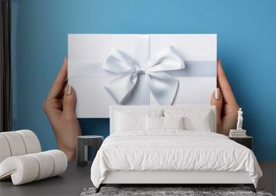 Woman holding white present box with vivid silver ribbon bow as presenf for Christmas, fathers day, birthday on light blue background. Flat lay, copy space Wall mural