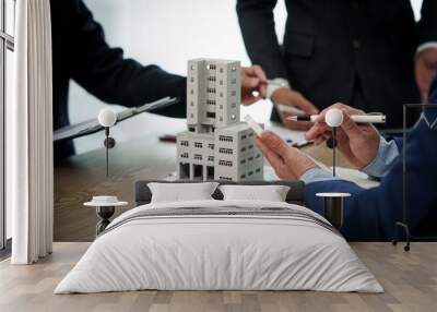 A professional businessman in a suit conducts a meeting at a desk while showcasing a model of a condominium or apartment building, discussing real estate development plans. Wall mural