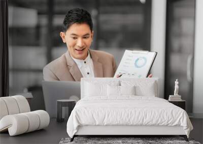 Asian professional male business person in beige suit and white shirt is working on laptop and holding pen, looking focused. Wall mural