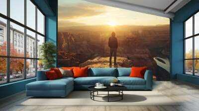 The image shows a person standing on a cliff, looking out at a vast canyon. The sun is setting, and the sky is ablaze with color. Wall mural