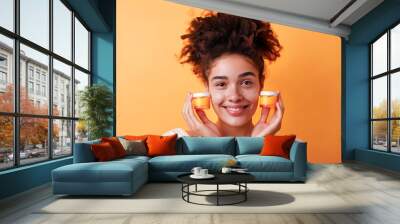 A beautiful woman with natural curly hair is smiling and holding two small yellow jars of face cream in her hands, copy space, advertising skin care Wall mural