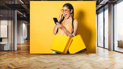 Beautiful young woman with shopping bags using her smart phone on yellow background.Shopaholic shopping Fashion. Wall mural