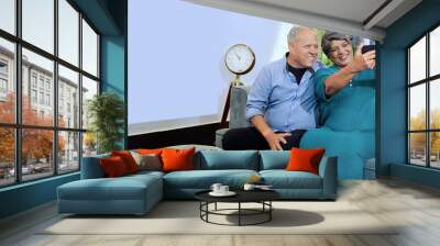 Indian happy old elder adult pair couple man lady sitting cozy sofa couch take funny selfie pose look phone cam indoor home aged older male female wife use mobile record video call enjoy fun joy Wall mural