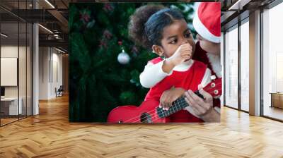 Happy a little child girl mixed-race Thai Afro-American with mom relaxing bonding and ukulele with a Christmas tree background. Loving black family cuddling fun and ukulele with family X mas. Wall mural