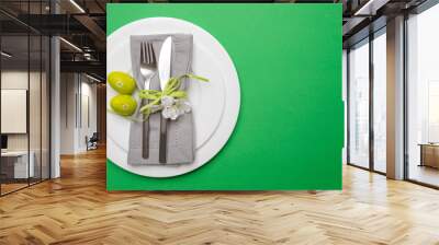 Easter eggs in a white plate, gray napkin and cutlery, green color background, top view Wall mural