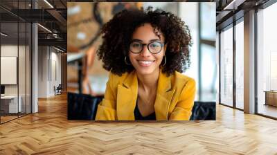 A young professional woman in a bright yellow blazer, smiling warmly while seated in a modern office environment. She wears stylish glasses and her curly hair frames her face beautifully Wall mural