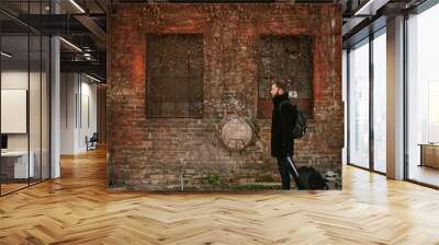 Photo taken from the side, in front of a brick wall. A guy with a beard, in a black coat, carries a backpack on his back and pulls his suitcase. Wall mural