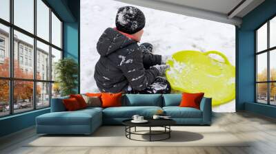 A boy of seven years old sitting on the snow and a green plastic saucer sled lying near him. Concept of winter activities, recreation and children's entertainment Wall mural