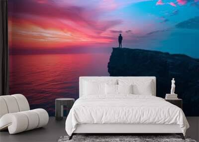 A dramatic seaside cliff at sunset, with the silhouette of a person standing at the edge, gazing out over the brightly colored horizon Wall mural