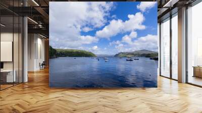 A serene landscape featuring a body of water with boats and buildings along the shore. The image captures a peaceful scene with a cloudy sky overhead.  Portree, Scotland, isle of skye Wall mural