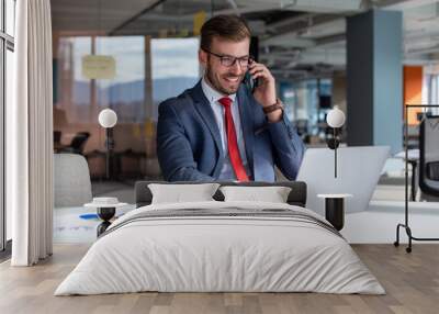 Portrait of a handsome businessman in eyeglasses talking on the phone with a client while sitting in the office. Wall mural