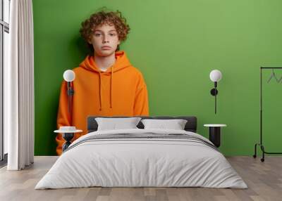 A relaxed teenage boy with curly hair standing against a green wall in an orange hoodie Wall mural