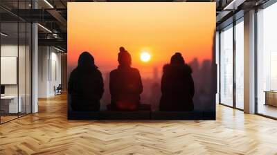 Three silhouetted friends in winter clothing sit on a ledge, watching a beautiful sunrise over a city skyline in winter. Wall mural