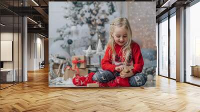 Little girl opening christmas present, looking at gift box with smile hild wearing Chrismas jumper posing on floor near fireplace and xmas tree. Wall mural