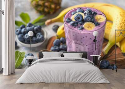 A close-up of a refreshing blueberry banana smoothie topped with blueberries and banana slices. The smoothie is in a glass and sitting on a wooden board with fresh fruit around it Wall mural