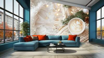 A warm cup of latte with latte art rests on a cozy knitted blanket, surrounded by Christmas lights and greenery Wall mural