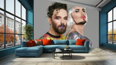 A transgender couple, 2 attractive male persons with beards and makeup on their faces, looking at the camera. The guy with a quiff touching the back of his partner in women's clothing and bob-cut wig. Wall mural