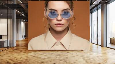 Cropped front view shot of lady with fair hair, wearing shirt. The girl with bun and wavy hair locks in oval-shaped glasses with glitter in blue lenses, is looking at camera on the brown background. Wall mural