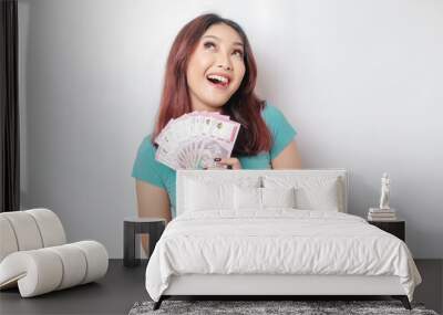 A happy young woman is wearing blue t-shirt and holding cash money in Indonesian rupiah isolated by white background Wall mural