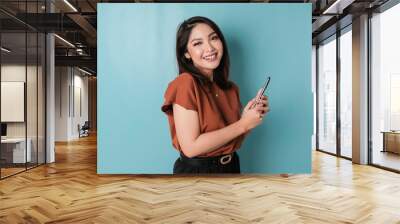 A portrait of a happy Asian woman is smiling and holding his smartphone wearing brown shirt Wall mural