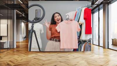 Cheerful young Asian woman doing live stream online shopping presenting pink shirt while showing okay finger gesture. Wall mural