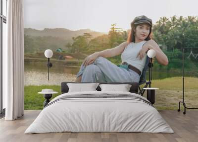 Elegant young Asian woman is sitting in a lawn chair by a lake. She is wearing a white sleeveless top and black hat. Wall mural