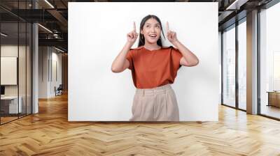 Excited young Asian woman in a brown shirt and taupe pants pointing to the copy space above her, isolated by white background. Wall mural