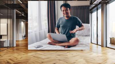 Happy Asian man is working with his laptop on his bed. Concept of freelancer successful lifestyle. Wall mural