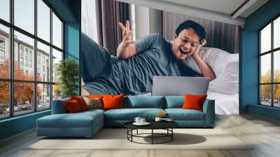 Happy Asian man is working with his laptop on his bed. Concept of freelancer successful lifestyle. Wall mural