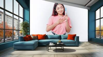 Happy mindful thankful young woman wearing pink t-shirt showing her pregnancy test, hand on chest smiling isolated on white background Wall mural