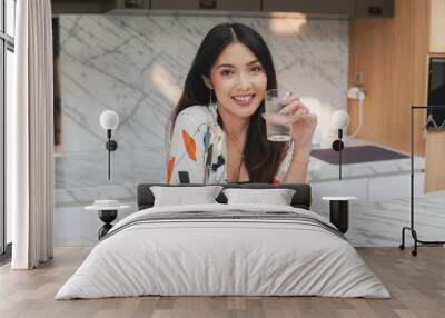 Joyful Asian woman is smiling and holding a glass of water in a kitchen island made from marble. Wall mural