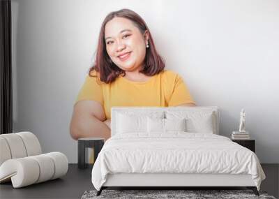 Portrait of a confident smiling big sized Asian woman wearing yellow shirt standing with arms folded and looking at the camera isolated over white background Wall mural