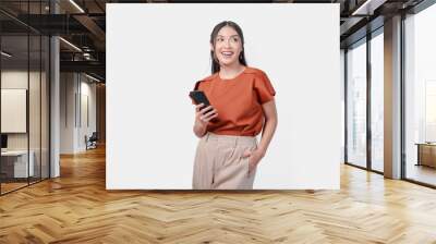 Smiling young Asian woman in a brown shirt is using her smartphone to look at social media, isolated by white background. Wall mural