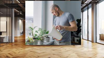 A young caucasian man watering houseplants on windowsill. Indoor gardening. Urban jungle. Father doing household chores. Wall mural