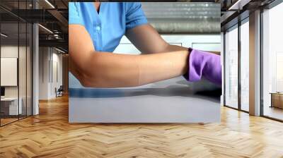 Woman in a blue shirt and purple gloves meticulously wiping a table with a rag, light grey background, professional color grading. Wall mural