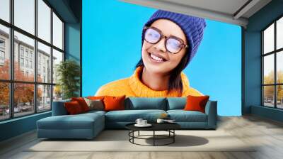 Portrait of happy woman in winter fashion with glasses, beanie and smile isolated on blue background. Style, happiness and face of gen z girl on studio backdrop with warm clothing for cold weather. Wall mural