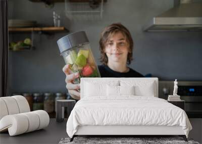 teen boy holding smoothie in focus with vegetables and fruit and smiling in background Wall mural