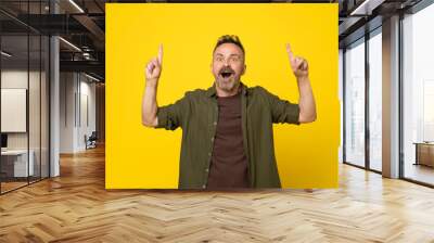 Astonished with open mouth handsome mature bristle man with two hands pointing up to empty space above isolated yellow background dressed in green shirt and brown t-shirt Wall mural