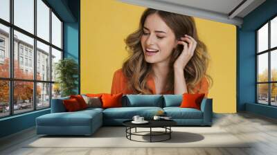 adorable woman in an orange attire touching her brown wavy hair. Wall mural