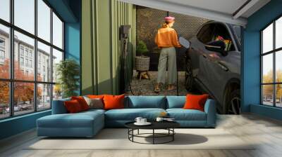 A woman in a stylish outfit charges her electric car at a modern home nestled in nature, highlighting a blend of sustainable living and contemporary lifestyle Wall mural