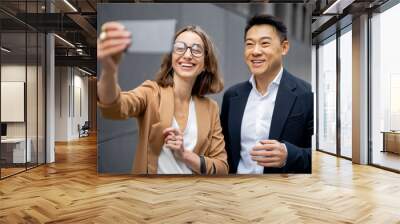 Asian businessman and caucasian businesswoman taking selfie on smartphone on city street. Concept of business cooperation. Idea of modern successful business people. Man waving hand at camera Wall mural